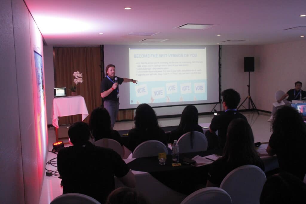 Bobby Macey delivering a presentation at the Kajae Summit in Cancún 2025, pointing to a slide about becoming the best version of yourself while speaking to the audience.