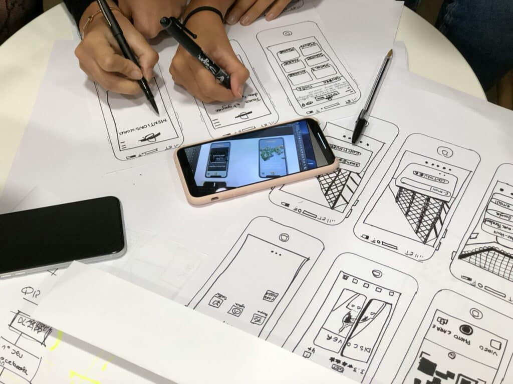Two people sketching mobile app wireframes on paper, surrounded by smartphones showing interface designs.